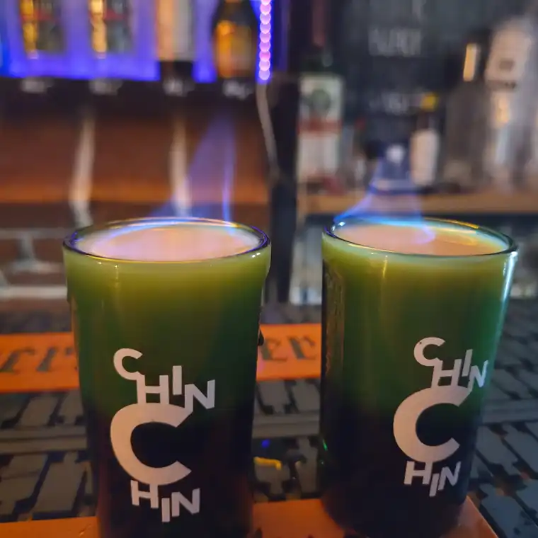 Dwa płonące zielone shoty w szklankach z napisem CHIN CHIN na barze